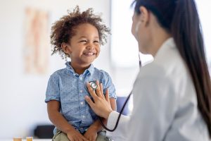 A female doctor of Asian decent listens to her patients heart with her stethoscope.  The little boy is sitting up on the exam table smiling back at his doctor as she holds the instrument to his chest.